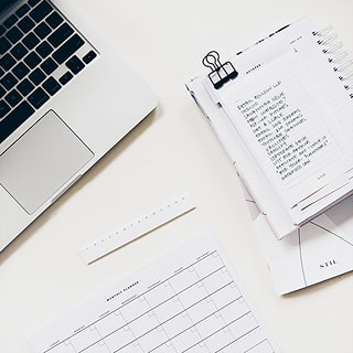 デスクに置かれたノートパソコン、定規、手書きのメモが挟まれたノートやスケジュール帳